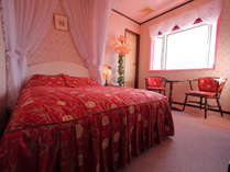 大きな出窓とベッドを囲む白のレースカーテンと花柄の赤が鮮やかなフリルのバッドカバーで優雅さを演出。